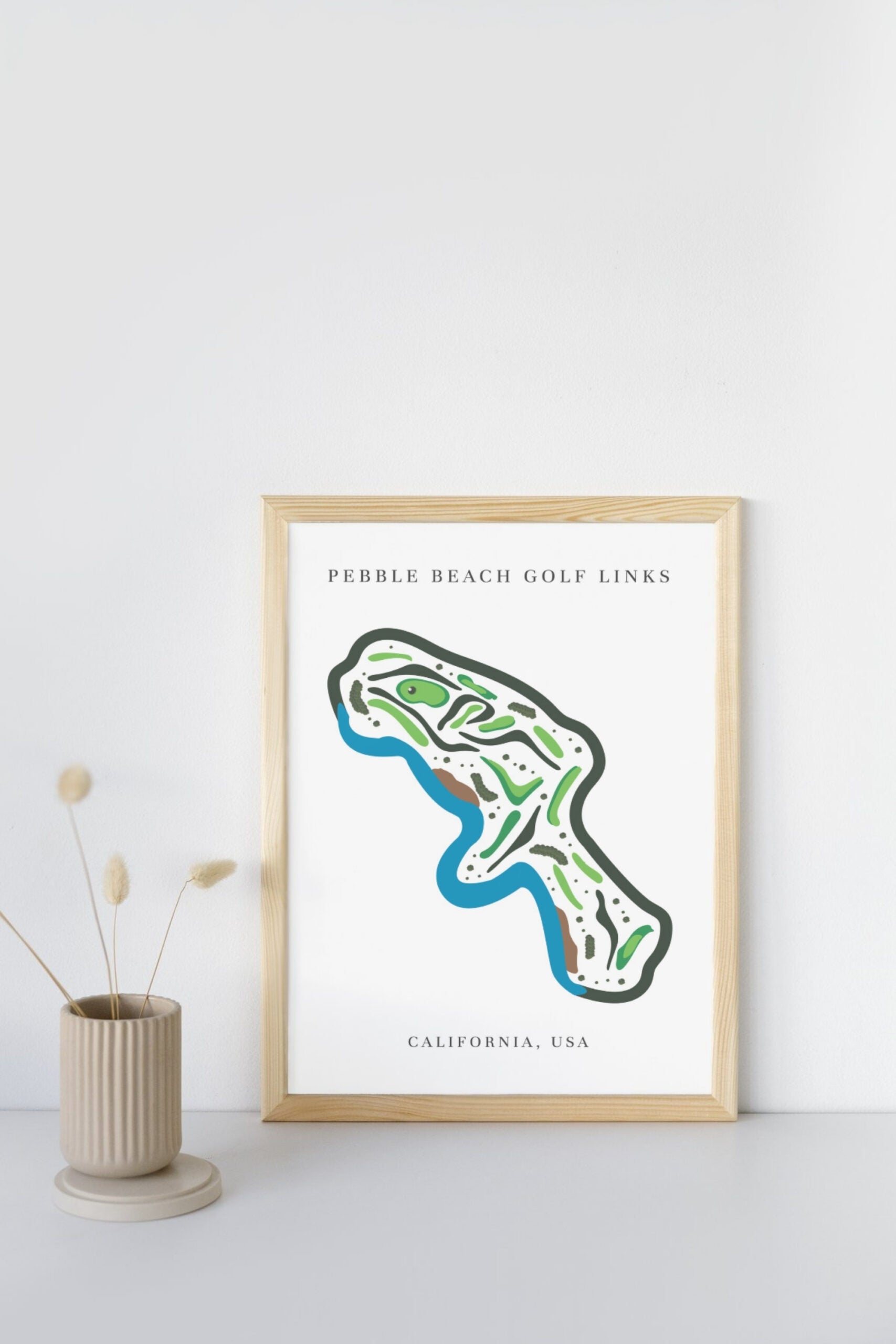 Framed map of Pebble Beach Golf Links on a white wall with a vase and plant in the foreground.
